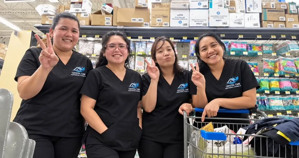 Four Crystal Care team members in uniform smiling and making peace signs in a store aisle with a cart of cleaning supplies
